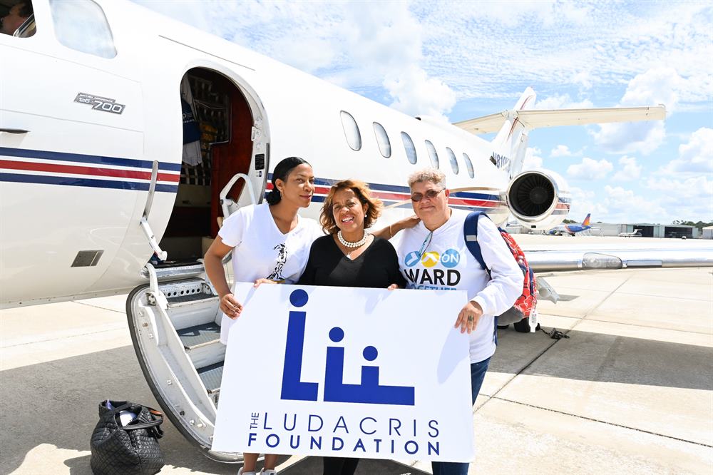 Roberta Shields and two women holding a Ludacris Foundation sign in front of a jet.