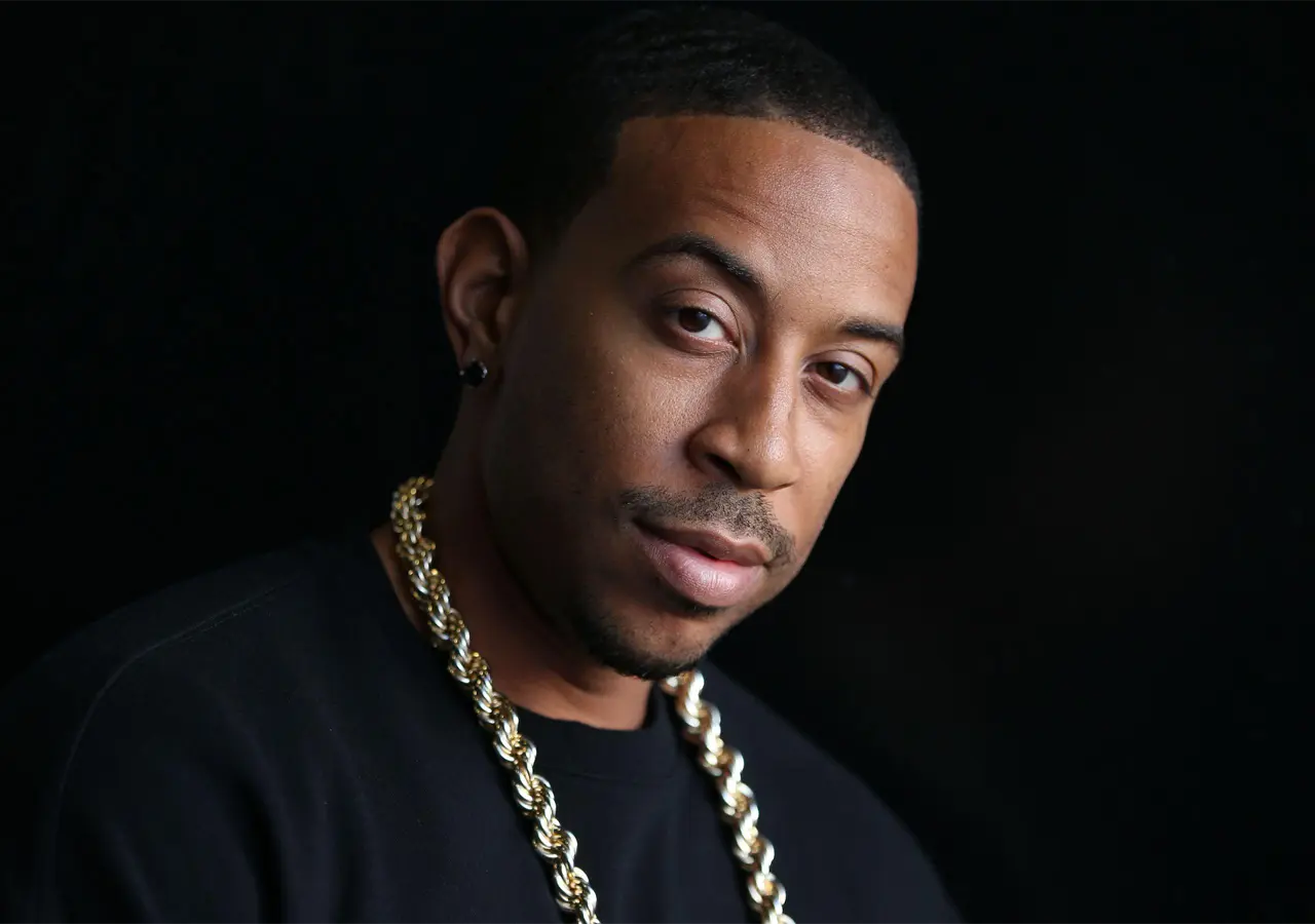 A close-up image of Chris 'Ludacris' Bridges wearing a black shirt and gold chain against a black background.