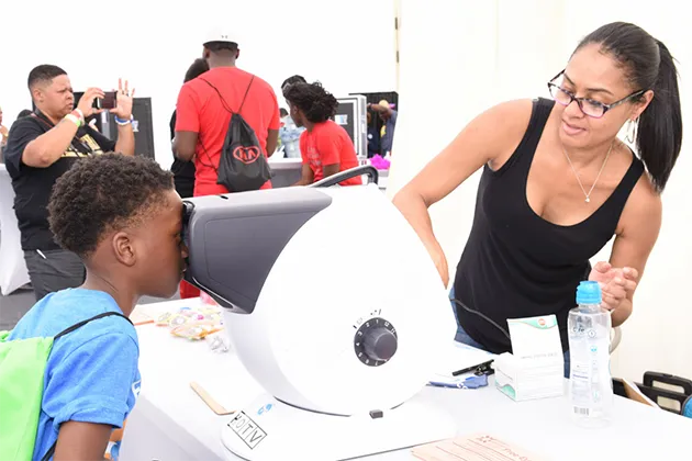 A child wearing a blue t-shirt and green backpack having his vision tested by a woman wearing a black shirt.