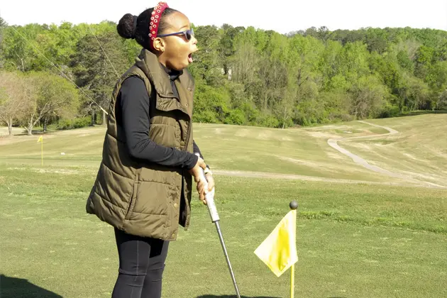 An excited young person holding a golf club stands on a golf course next to a yellow flag.
